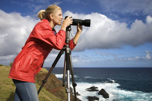 Caucasian mid-adult woman looking through camera on tripod on cliff overlooking ocean in Maui, Hawaii.