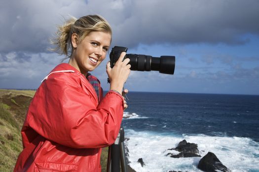 Caucasian mid-adult woman holding camera on tripod, looking at viewer, on cliff overlooking ocean in Maui, Hawaii.