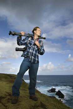 Caucasian mid-adult man standing with camera and tripod over his shoulder on cliff overlooking ocean in Maui, Hawaii.