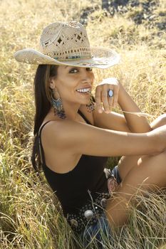 Mid-adult Caucasian woman sitting in grass wearing straw cowboy hat chewing on blade of grass.