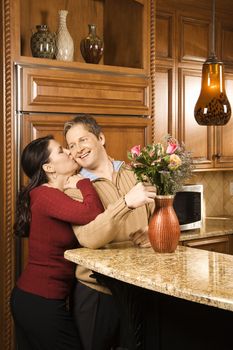 Caucasian woman kissing Caucasian man on cheek as he arranges flowers in vase in kitchen.