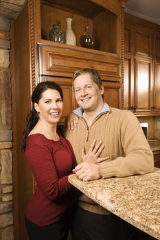 Caucasian woman and Caucasian man with arms around each other leaning on kitchen counter and looking at viewer.