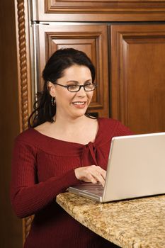 Caucasian woman typing on laptop in kitchen and smiling.
