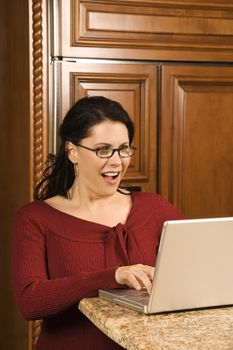 Caucasian woman typing on laptop in kitchen and looking at monitor with shocked expression.