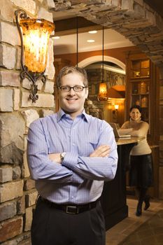 Caucasian man standing with arms crossed looking at viewer and smiling with Caucasian women in background with laptop.