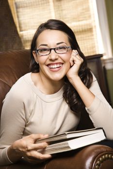 Caucasian/Hispanic young woman sitting in leather chair holding book and smiling at viewer.