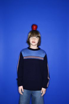 Portrait of Caucasian boy with an apple on his head standing against blue background.