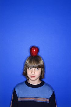 Head and shoulder portrait of Caucasian boy with apple on his head standing against blue background.