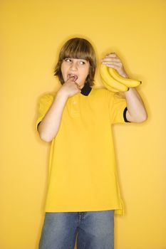 Portrait of Caucasian boy holding bunch of bananas and gesturing with finger in mouth that they are gross standing against yellow background.