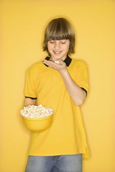 Portrait of Caucasian boy smiling holding bowl of popcorn in one hand and bringing some popcorn to mouth standing against yellow background.