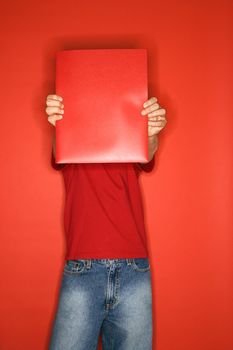 Portait of Caucasian boy covering his face with red folder standing against red background.