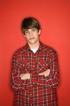 Portrait of Caucasian teen boy with arms crossed standing against red background wearing flannel shirt.