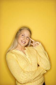 Portrait of Caucasian blond teen girl pointing at her head standing against yellow background.