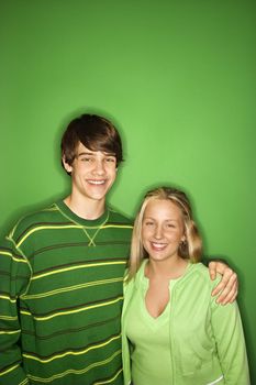 Portrait of Caucasian teen boy and girl standing against green background smiling with arms around eachother.