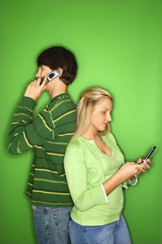 Portrait of Caucasian teen boy and girl on cellphones standing with backs to eachother against green background.