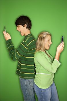 Portrait of Caucasian teen boy and girl on cellphones standing with backs to eachother against green background.