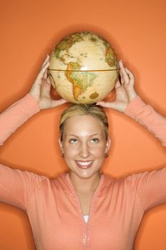 Portrait of Caucasian teen girl holding globe on her head smiling standing against orange background.