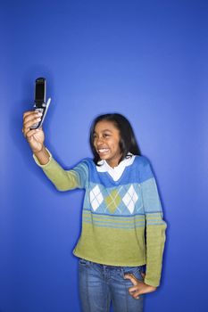 Portrait of African-American teen girl taking photo with camera phone standing in front of blue background.