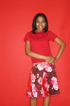 Portrait of smiling African-American teen girl with hands on her hips standing in front of red background.