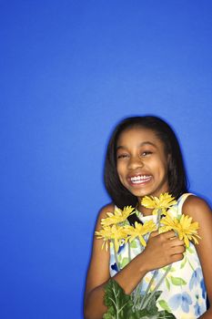 Portrait of African-American teen girl holding a bouquet of daisies and smiling against blue background.