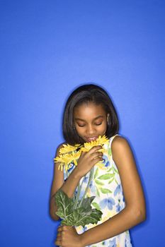 Portrait of African-American teen girl smelling a bouquet of daisies standing against a blue background.