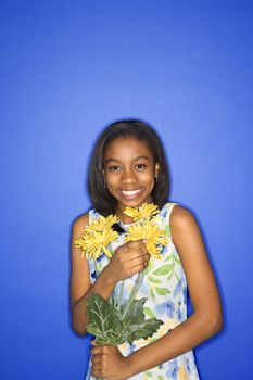Portrait of African-American teen girl holding a bouquet of daisies and smiling in front of blue background.