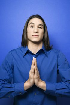 Portrait of Asian-American teen boy with hands pressed together in prayer position standing against blue background.