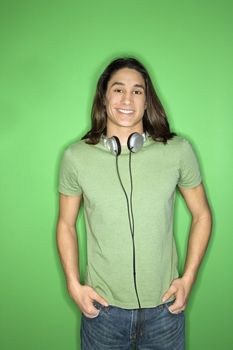 Asian-American teen boy smiling with headphones around neck and hands in pockets.