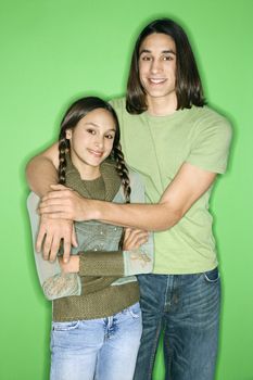 Portrait of Asian-American girl and teen boy standing with arms around eachother smiling against green background.