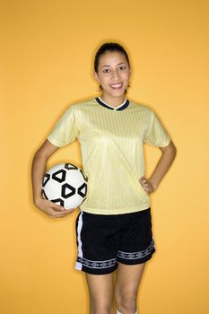 Portrait of smiling Multi-racial teen girl holding soccer ball at hip standing against yellow background.