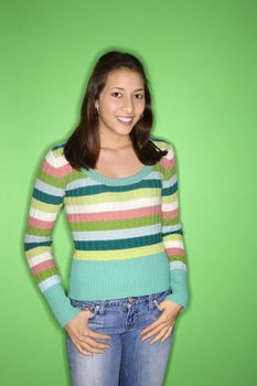 Portrait of smiling Multi-racial teen girl with thumbs in pockets standing against green background.