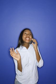 Portrait of smiling African-American teen girl looking up and talking on cellphone standing against blue background.