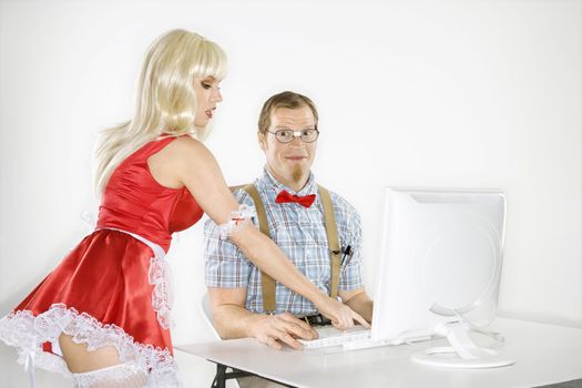 Caucasian young man smiling dressed like nerd sitting at computer looking at viewer with Caucasian young blonde woman in sexy french maid outfit bending over and pointing.