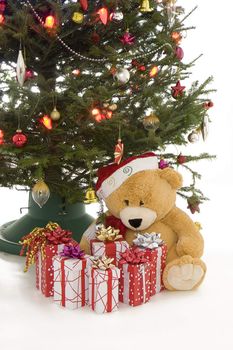 christmas tree with present in a red and white with a teddy bear sitting beside it