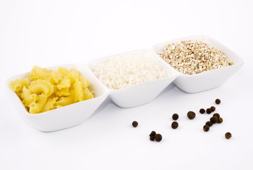 Three porceline bowls filled with pasta, groats and rice, on white background