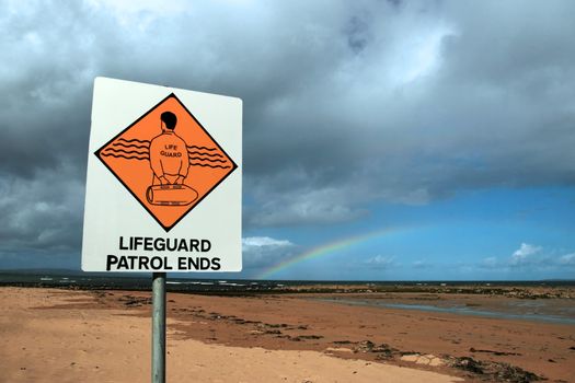 a sign warning that there is no lifeguard patrol beyond this sign with a beautiful rainbow in the background
