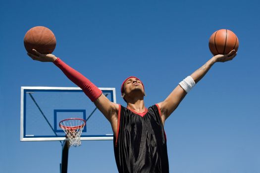 A young athletic build basketball player holding up two basketballs in the air.