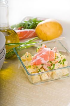 fresh home made parma ham and potato salad,with raw ingredients around with bowls and dishware on a table