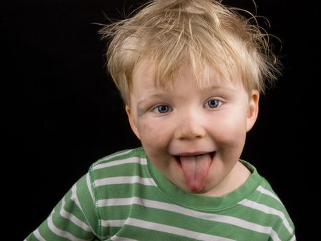 Little boy showing his toungue on black background. Boy have blue eyes, blond hair and a bit of dirt on face