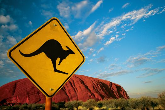 Kangaroo warning sign in front of Ayers Rock early in the morning as the sun turns it bright red.
