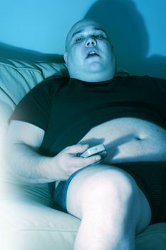 Lazy overweight male sitting on a couch watching television.  Harsh blue lighting from television with slow shutter speed to create TV watching atmosphere. Selective focus on the eyes.
