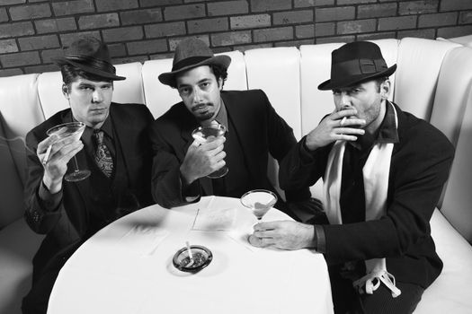 Three Caucasian prime adult males in retro suits sitting at table with cocktails looking at viewer.