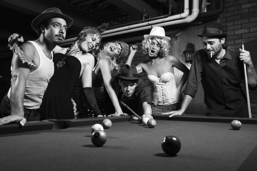 Group of Caucasian prime adult retro males and females trying to distract man as he takes pool shot.