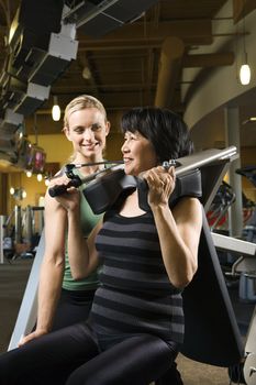 Prime adult Caucasian female trainer helping Asian mature adult female on exercise machine.