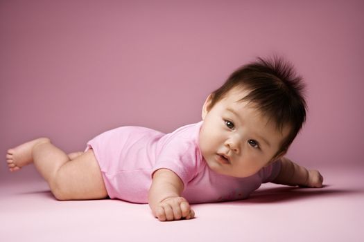 Asian baby lying on stomach looking at viewer holding arms out to side.