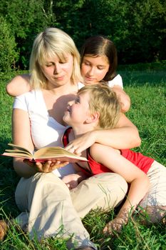 The young woman sits on a grass and reads the book to the children