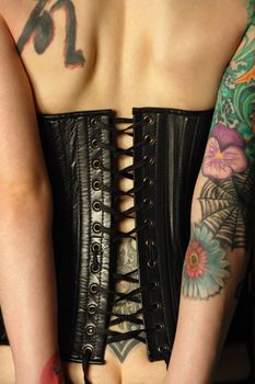 A young slim women with arm and back tattoos dressed in a black leather corset.  Focus is on the middle of corset.  
