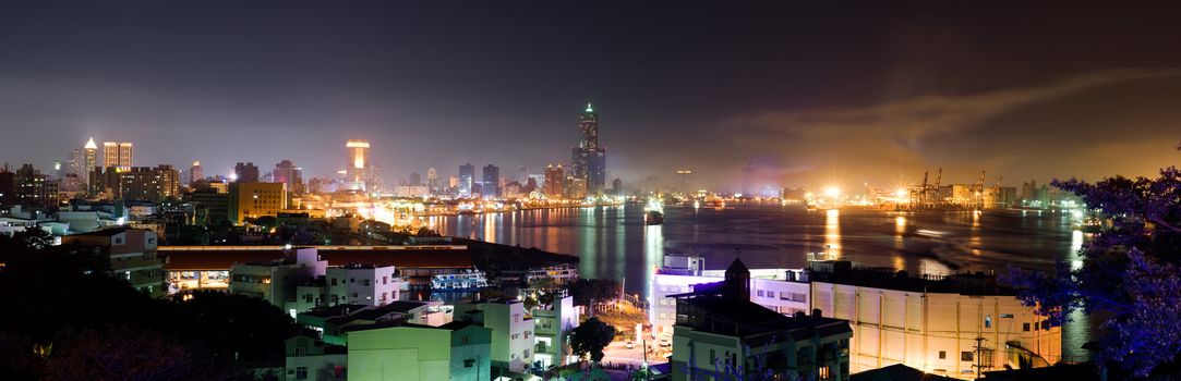 Color panoramic cityscape in night with boats in harbor of ocean and buildings of skyscraper in Kaohsiung, Taiwan.