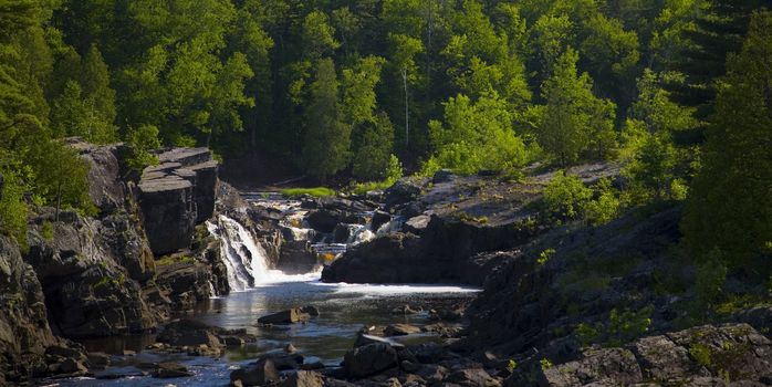 Water steps and falls down stone from a green forest into a blue river in Northern Minnesota.