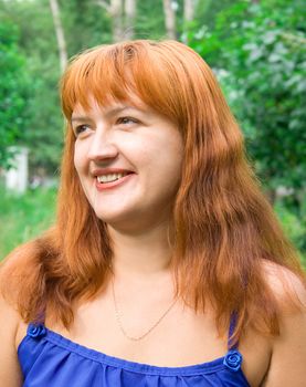 Portrait of the smiling woman with red hair in dark blue clothes against green plants